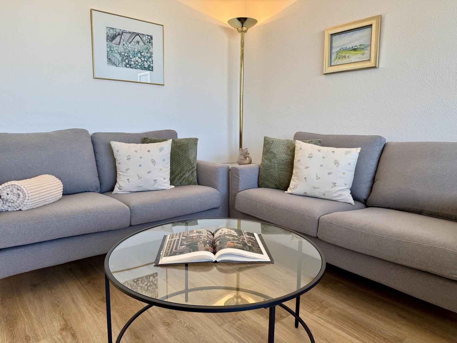 Zwei graue Sofas um einen runden Glas-Tisch mit offenem Buch.