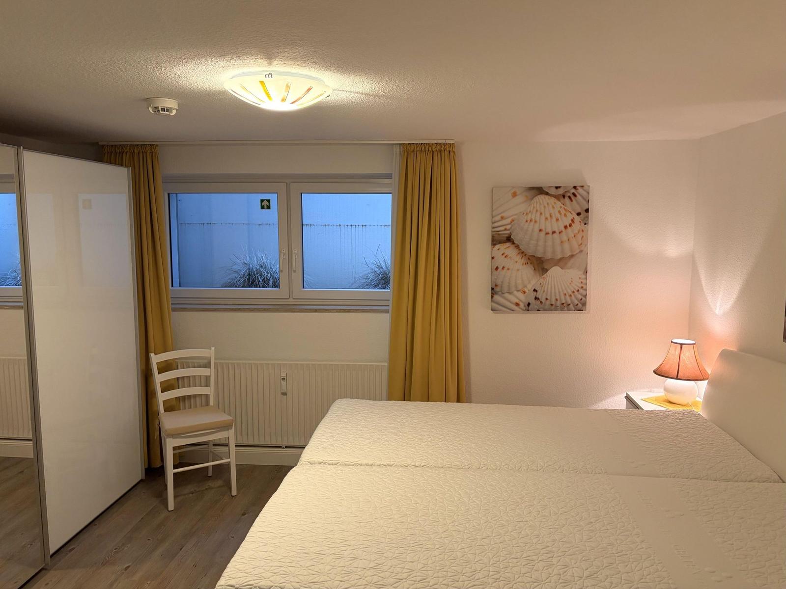 Schlafzimmer mit Bett, Nachttisch, Lampe und Fenster mit gelben Vorhängen.