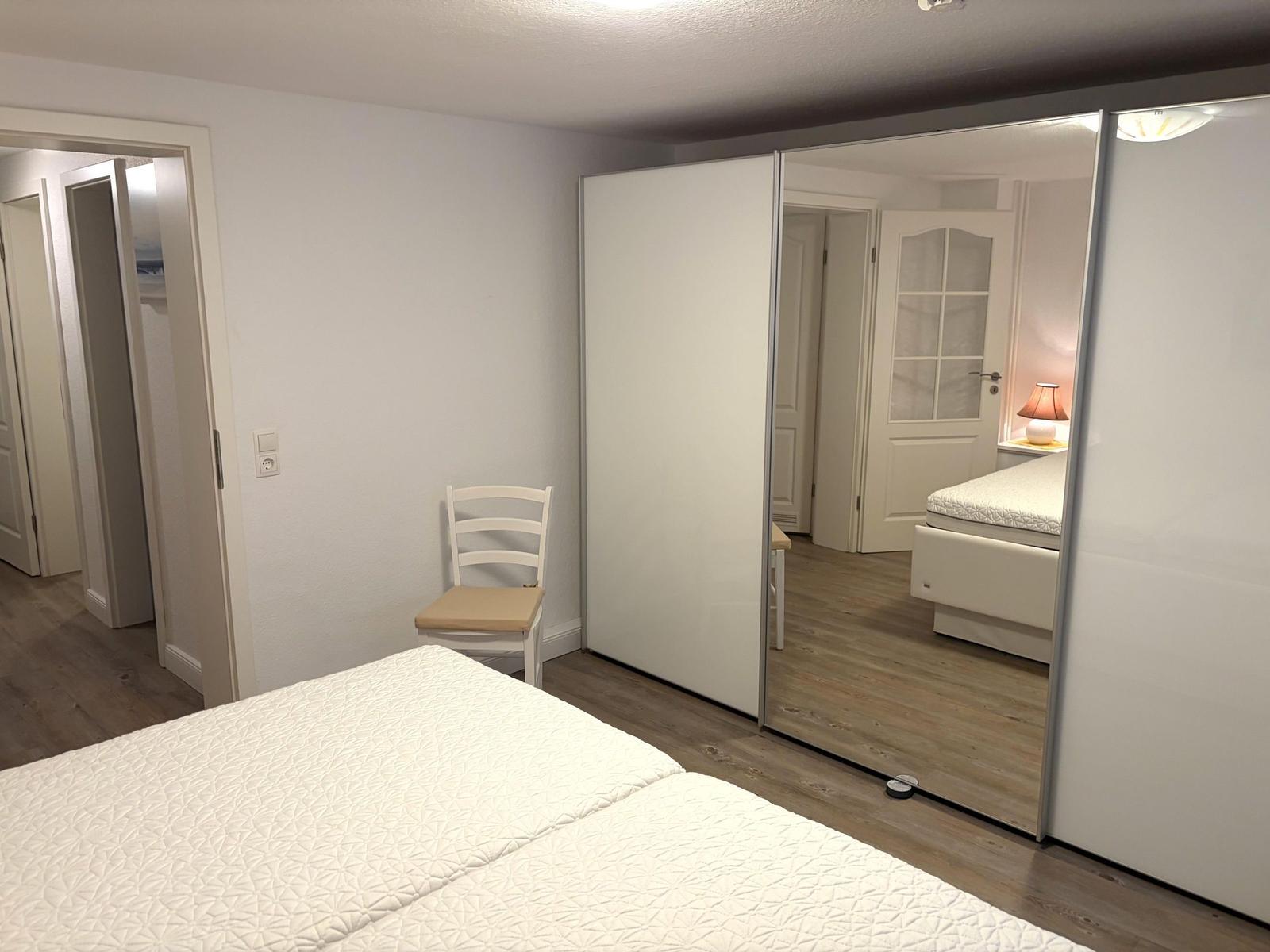 Schlafzimmer mit Bett, Stuhl und Spiegelschrank. Holzdielenboden und Türen im Hintergrund.