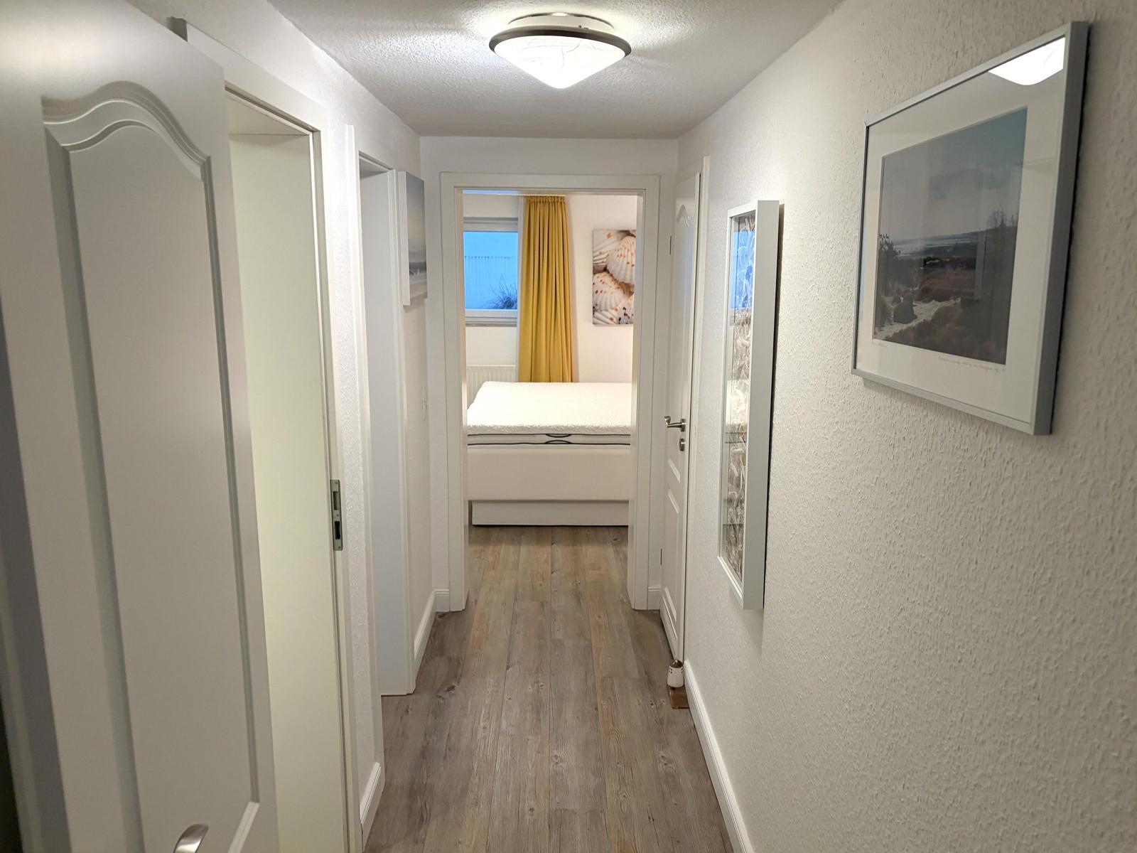 Flur mit Holzboden und weißen Wänden. Im Hintergrund ist ein Schlafzimmer mit Bett und Vorhang sichtbar.