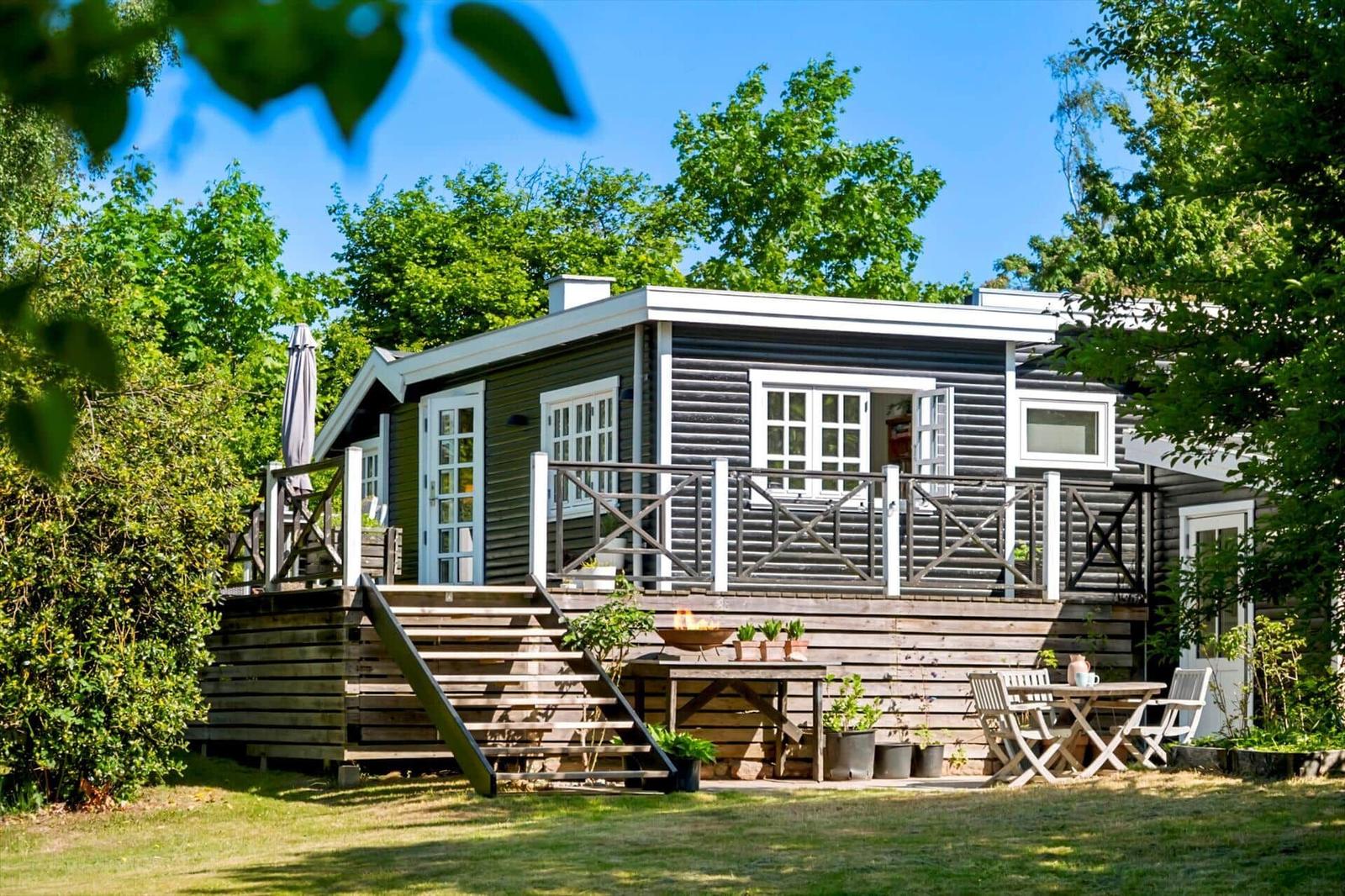 Wooden house with terrace, garden furniture, and barbecue area. Surrounded by trees under blue sky.