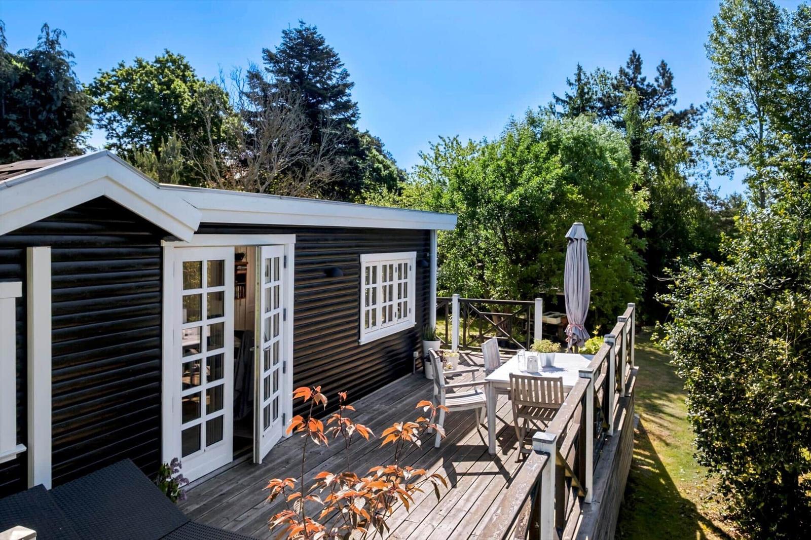 Black wooden house with terrace, table, chairs, and umbrella. Surrounded by trees and greenery.