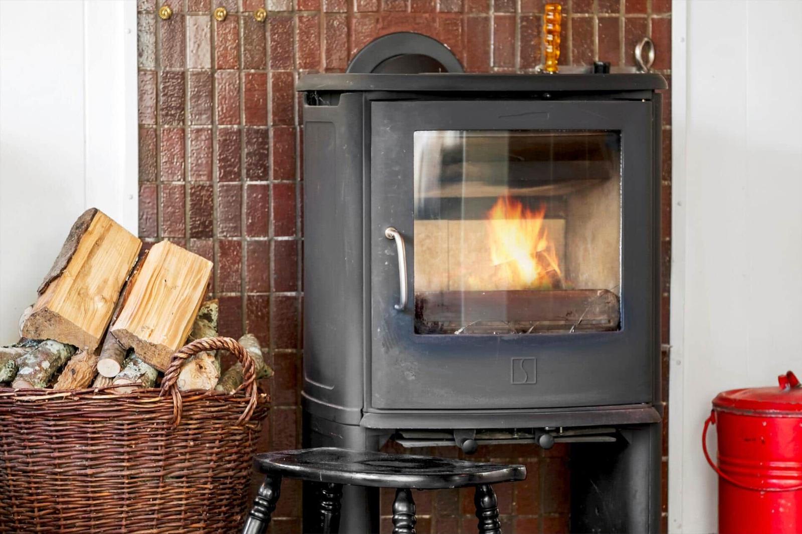 A wood stove with a burning fire, next to a basket of firewood and a red bucket.