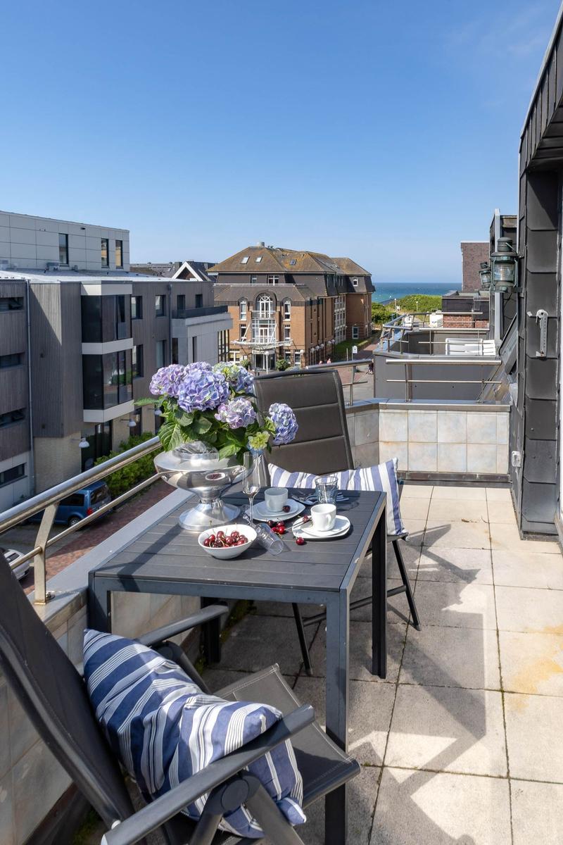 Terrasse mit Tisch, Stühlen und Blumen. Blick auf Gebäude und Meer.