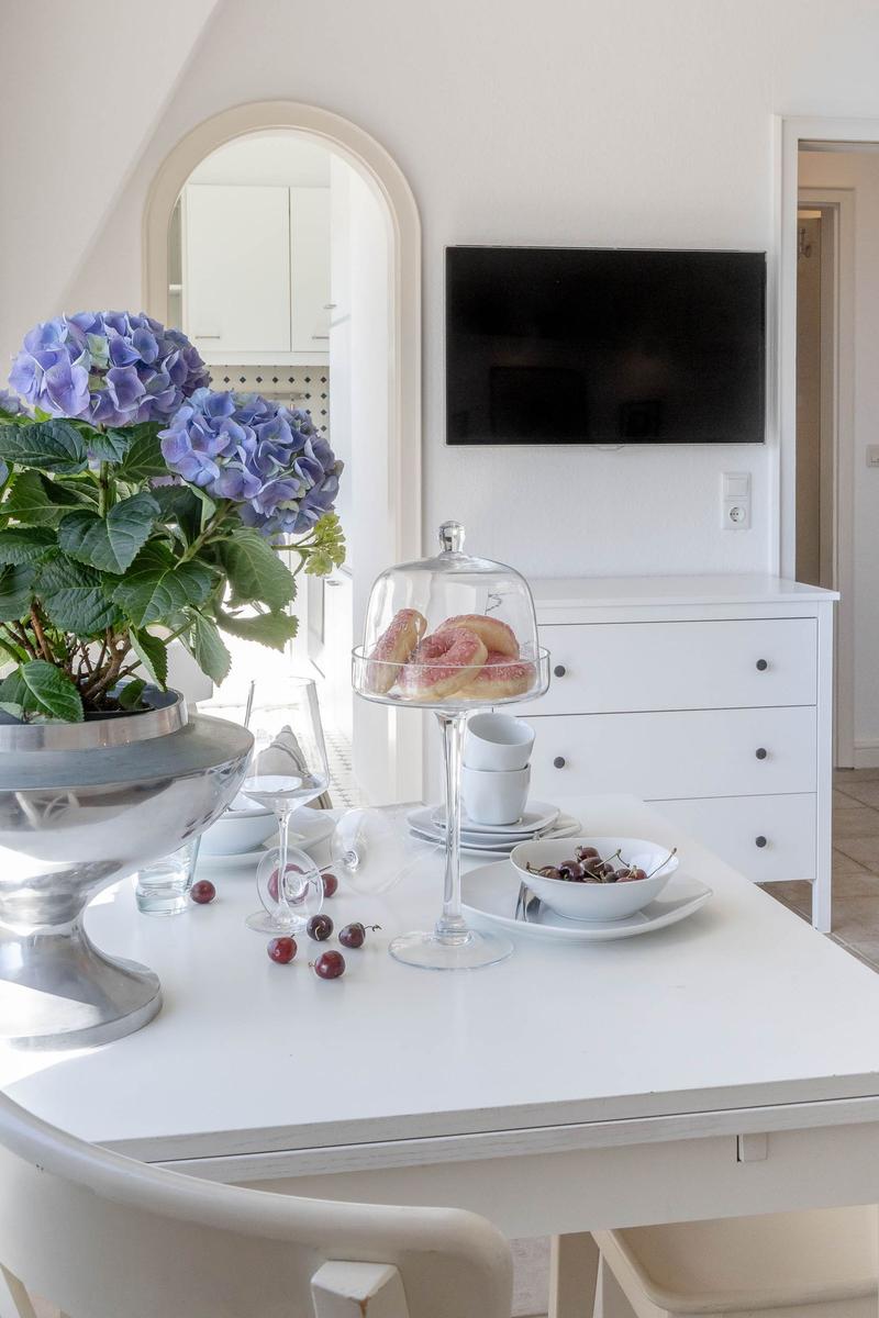 Tisch mit Blumen, Donuts und Kirschen. Hintergrund: Fernseher und Schrank.