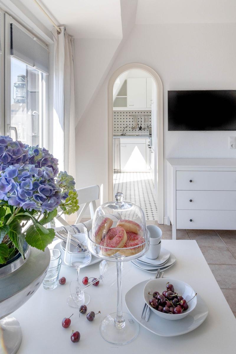 Tisch mit Donuts, Kirschen und Blumen vor Fenster. Küche im Hintergrund.