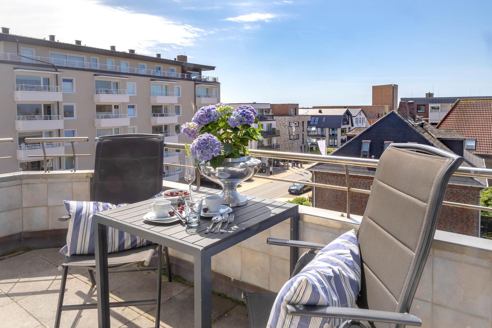 Terrasse mit Tisch, zwei Stühlen und Blumen. Blick auf Wohngebäude und Straßen.