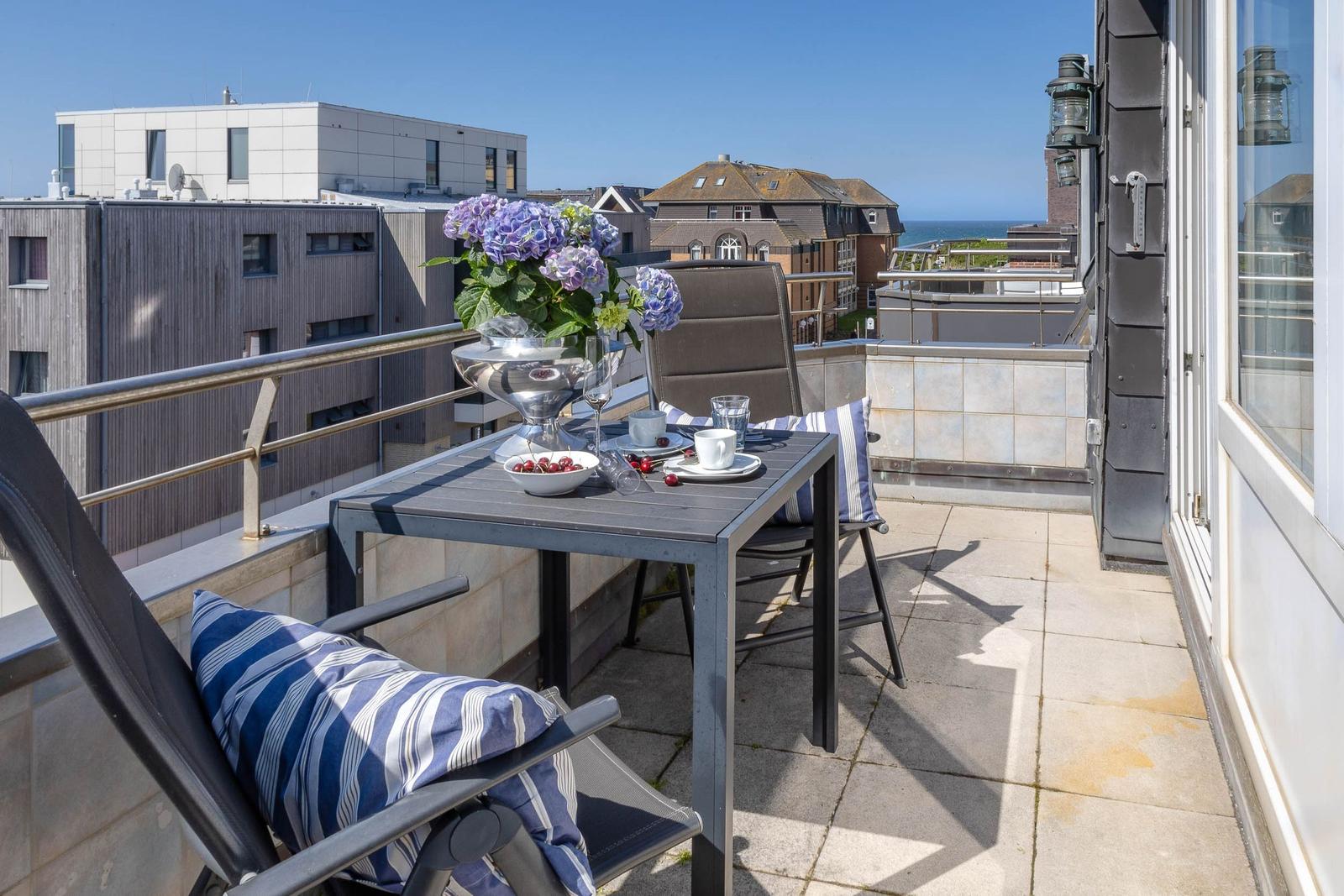 Terrasse mit Tisch, Stühlen und Blumen. Blick auf Gebäude und Meer.
