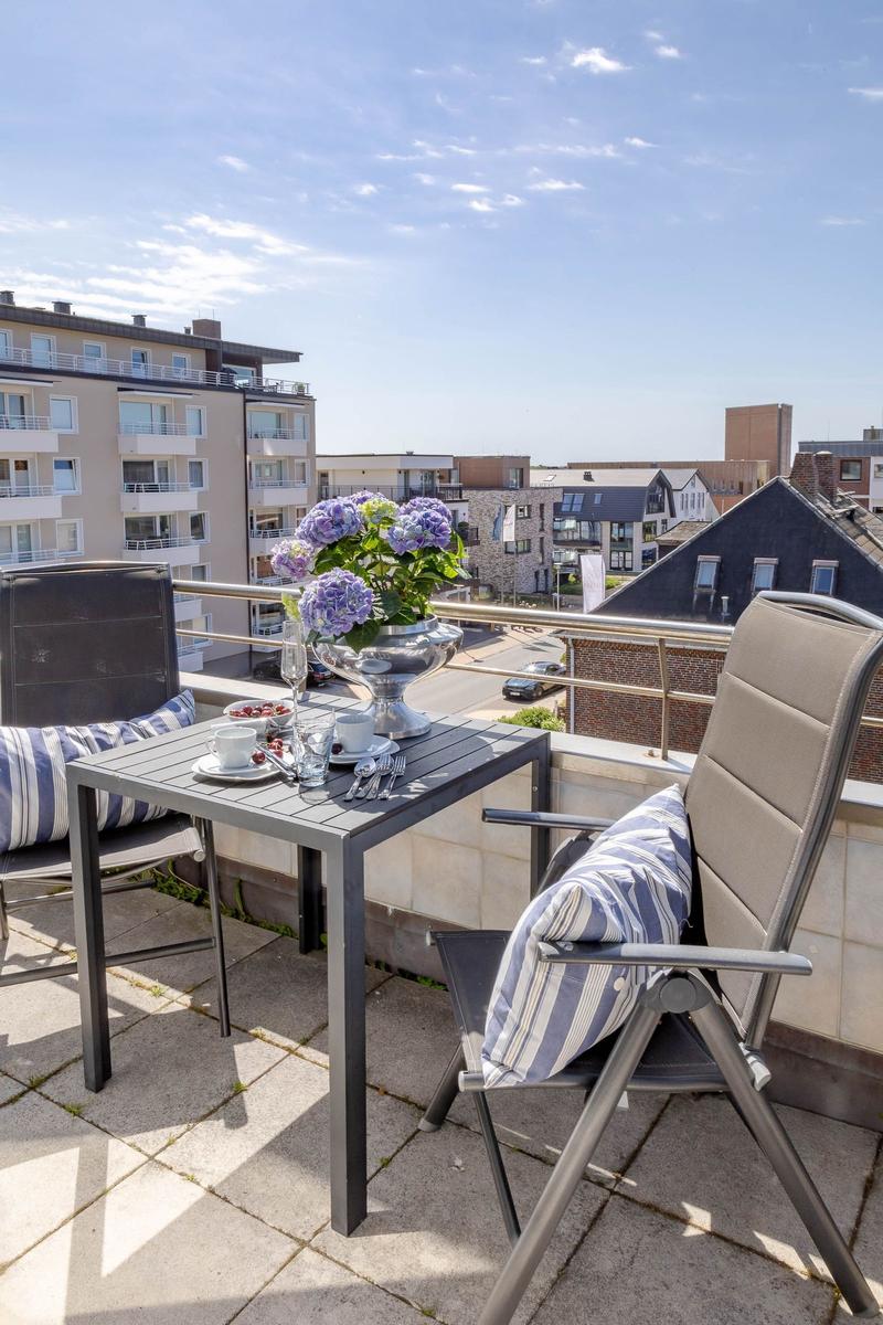Terrasse mit Tisch, zwei Stühlen und Blumen. Blick auf Wohngebiet unter blauem Himmel.