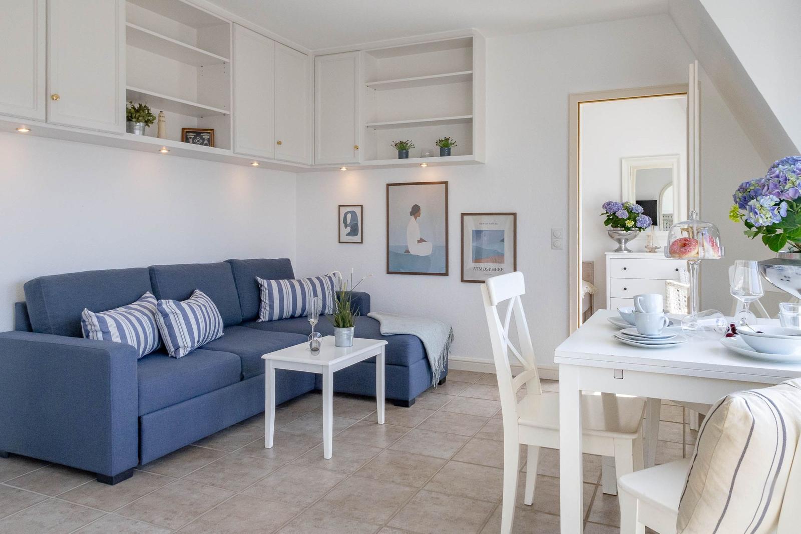 Wohn- und Essbereich mit blauem Sofa, weißem Tisch und Stühlen sowie Küchenregalen an der Wand.