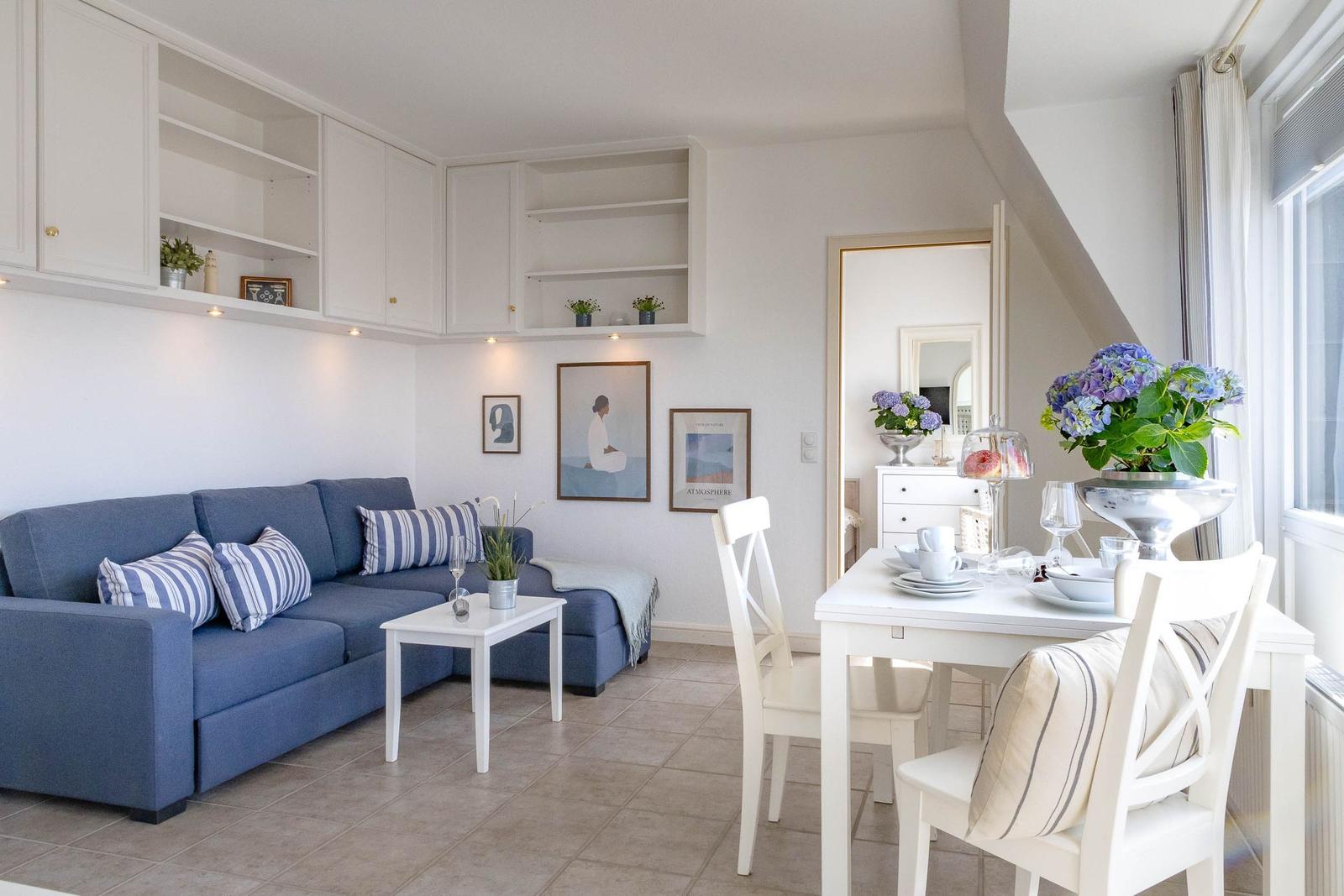 Wohn- und Essbereich mit blauem Sofa, weißem Tisch und Stühlen sowie Küchenregalen an der Wand.