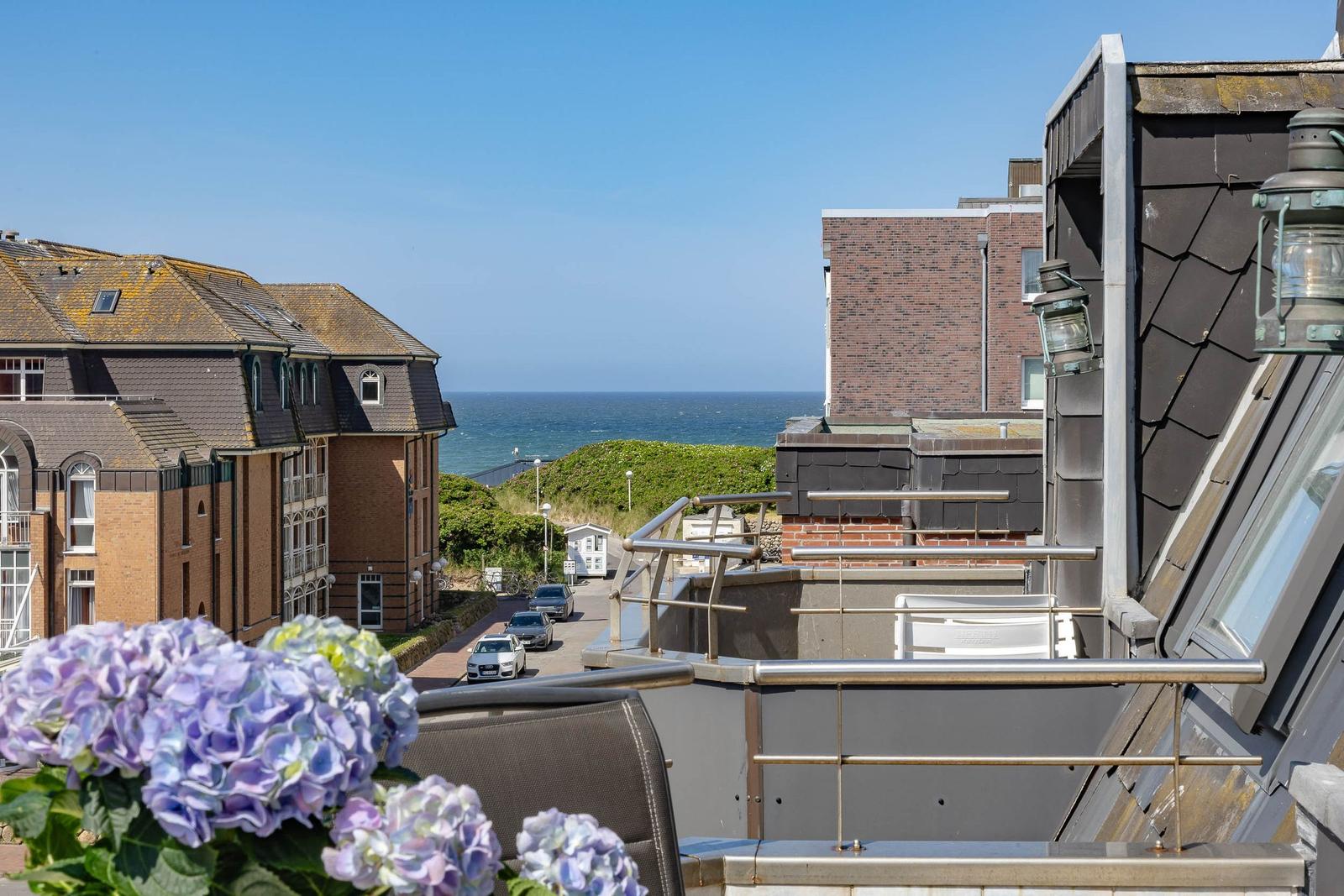 Blick von Balkon auf Straße, Häuser und Meer unter blauem Himmel mit Hortensien im Vordergrund.