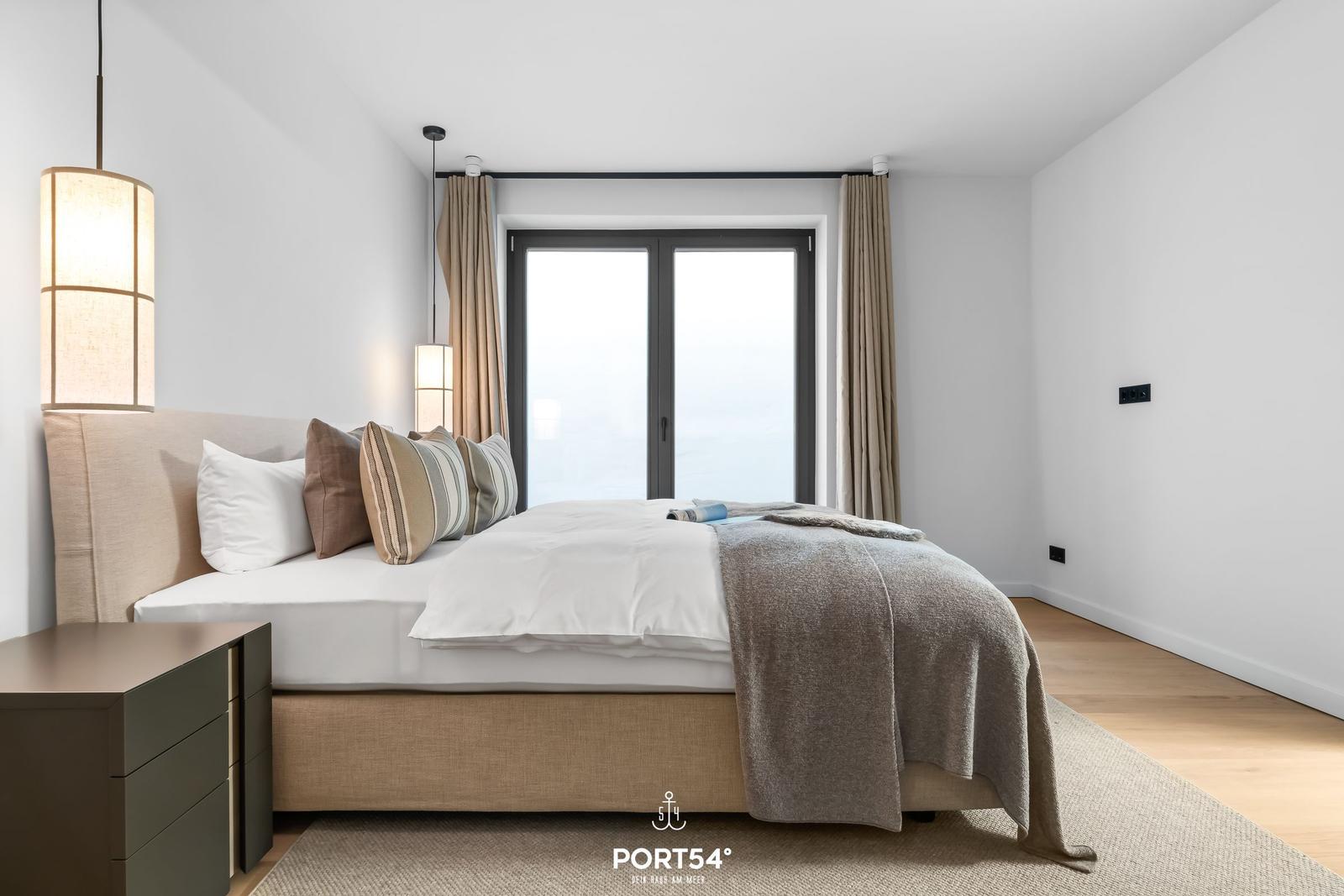 Schlafzimmer mit Bett, Nachttisch und Fenster. Holzboden und helle Wände.