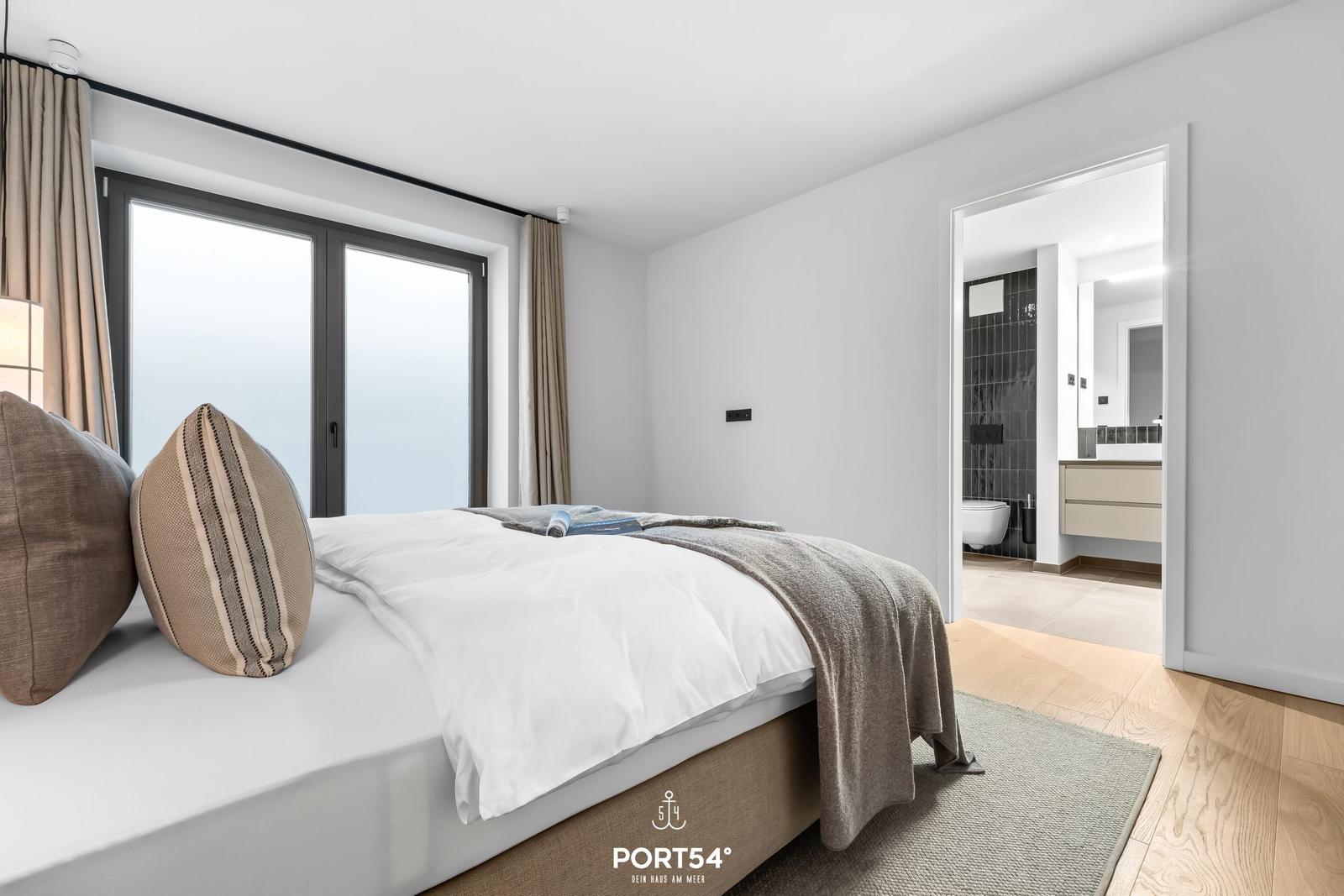 Schlafzimmer mit Bett, Fenster und Blick in das Badezimmer.