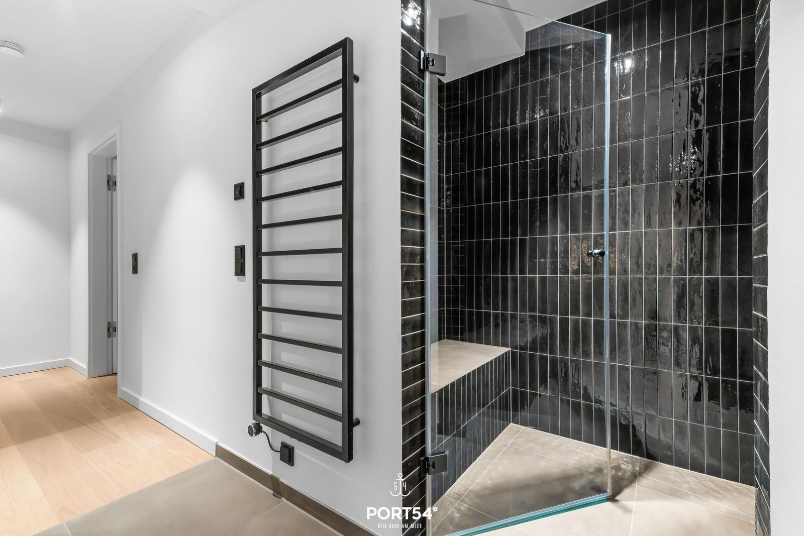 Dusche mit Glaswand und schwarzen Fliesen. Heizkörper an der Wand. Holzboden im Flur.