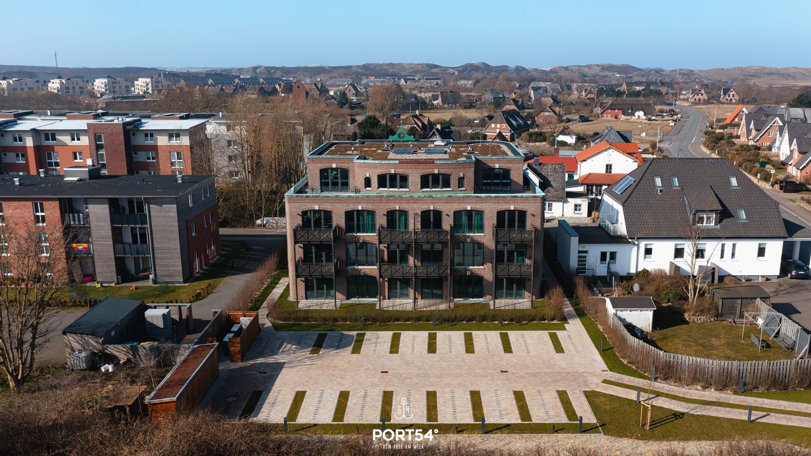 Moderne Wohnanlage mit Balkonen und gepflastertem Hof. Umgeben von Häusern und Hügeln.