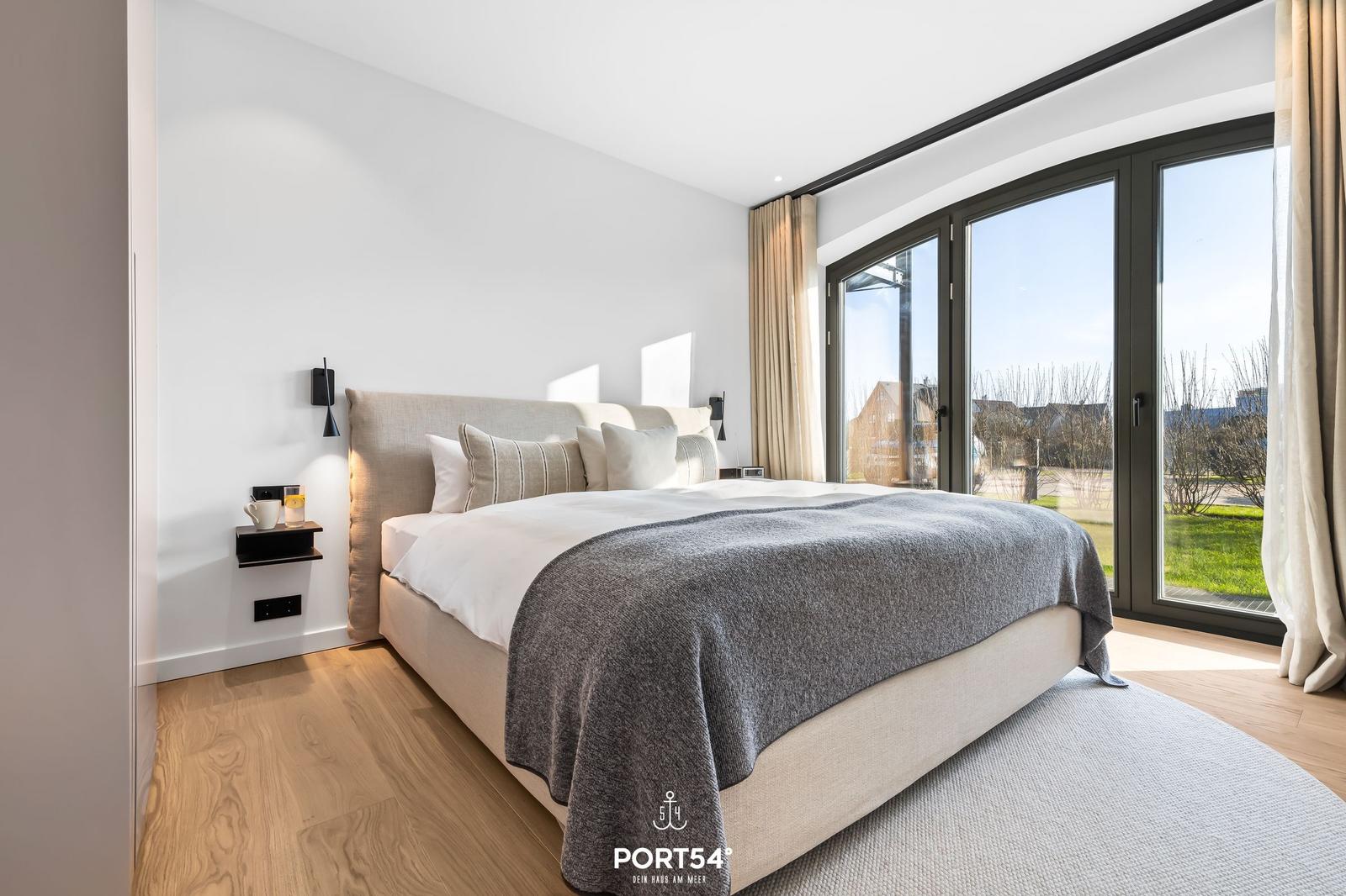 Schlafzimmer mit großem Bett, Holzboden und bodentiefen Fenstern mit Blick nach draußen.