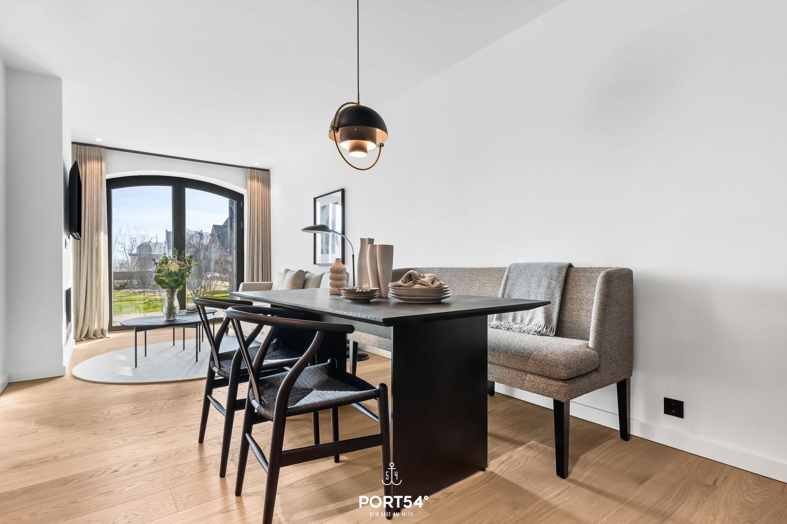 Essbereich mit Tisch, Stühlen und Sofa. Große Fenster mit Blick auf Außenbereich.