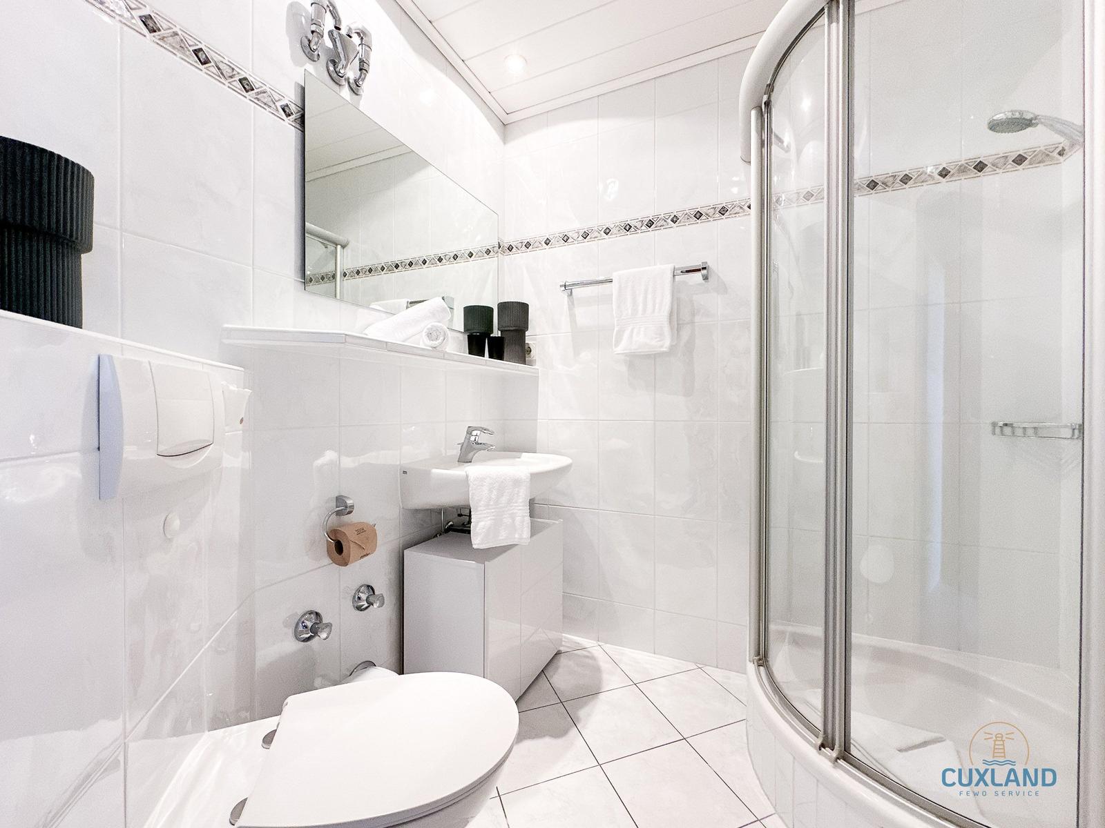 Badezimmer mit Dusche, Waschbecken und Toilette. Weiße Fliesen und Spiegel.