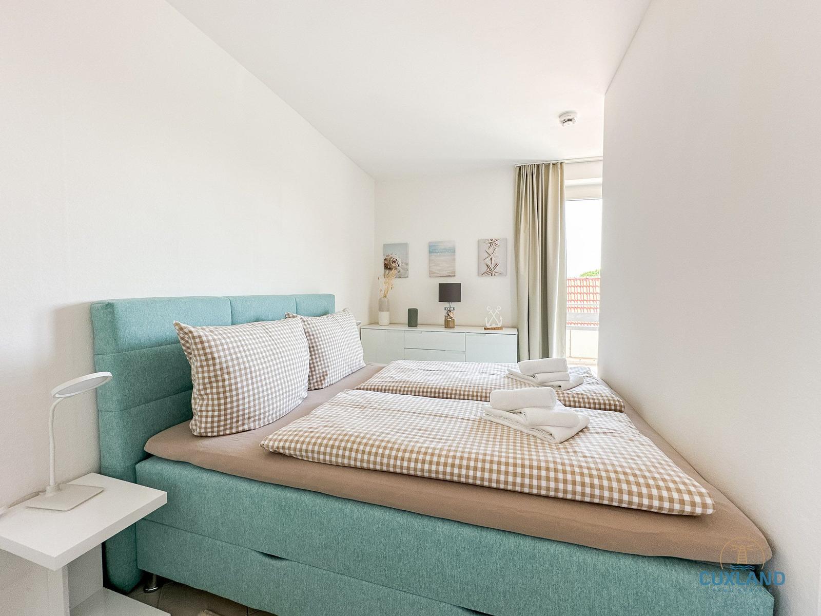 Schlafzimmer mit Bett, Nachttisch und Fenster. Wandbild und Vorhänge sind sichtbar.