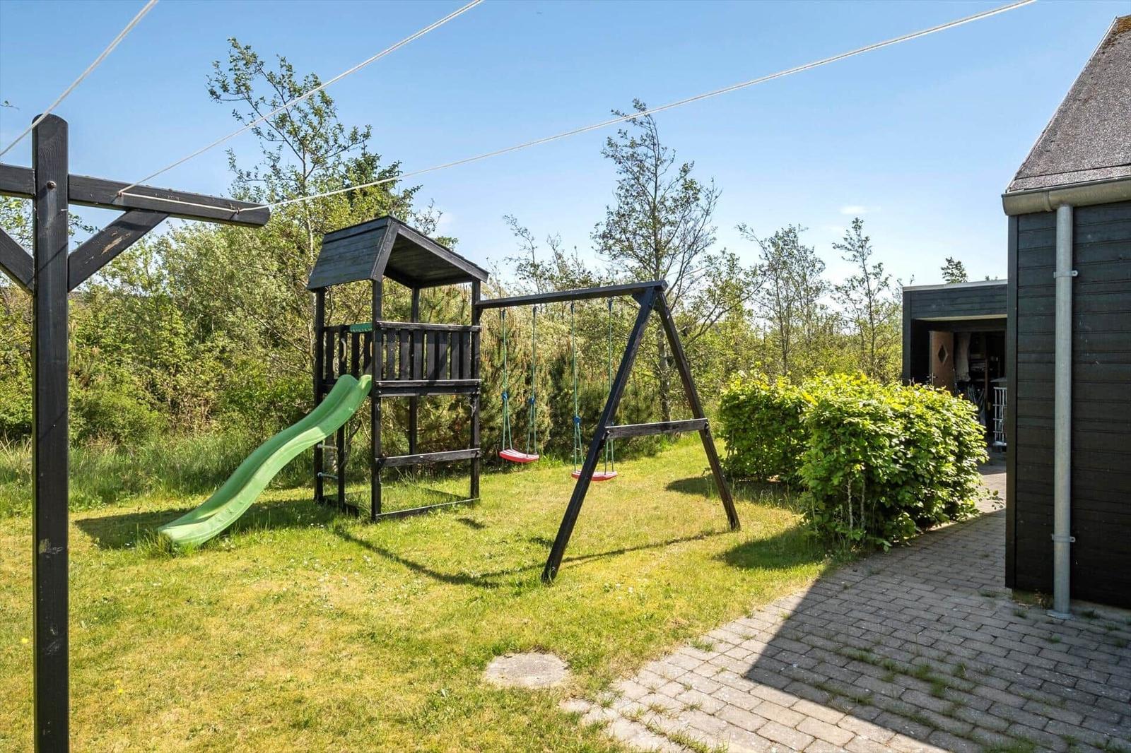 Backyard with playground, swing, and slide. Lawn and shrubs. Adjacent house with roof.