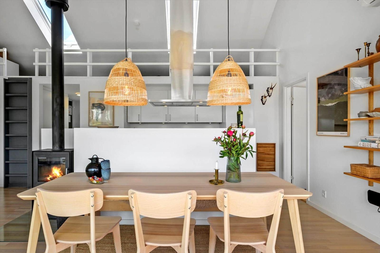 Dining area with wooden table, three chairs, and two pendant wicker lamps. Kitchen and fireplace in background.