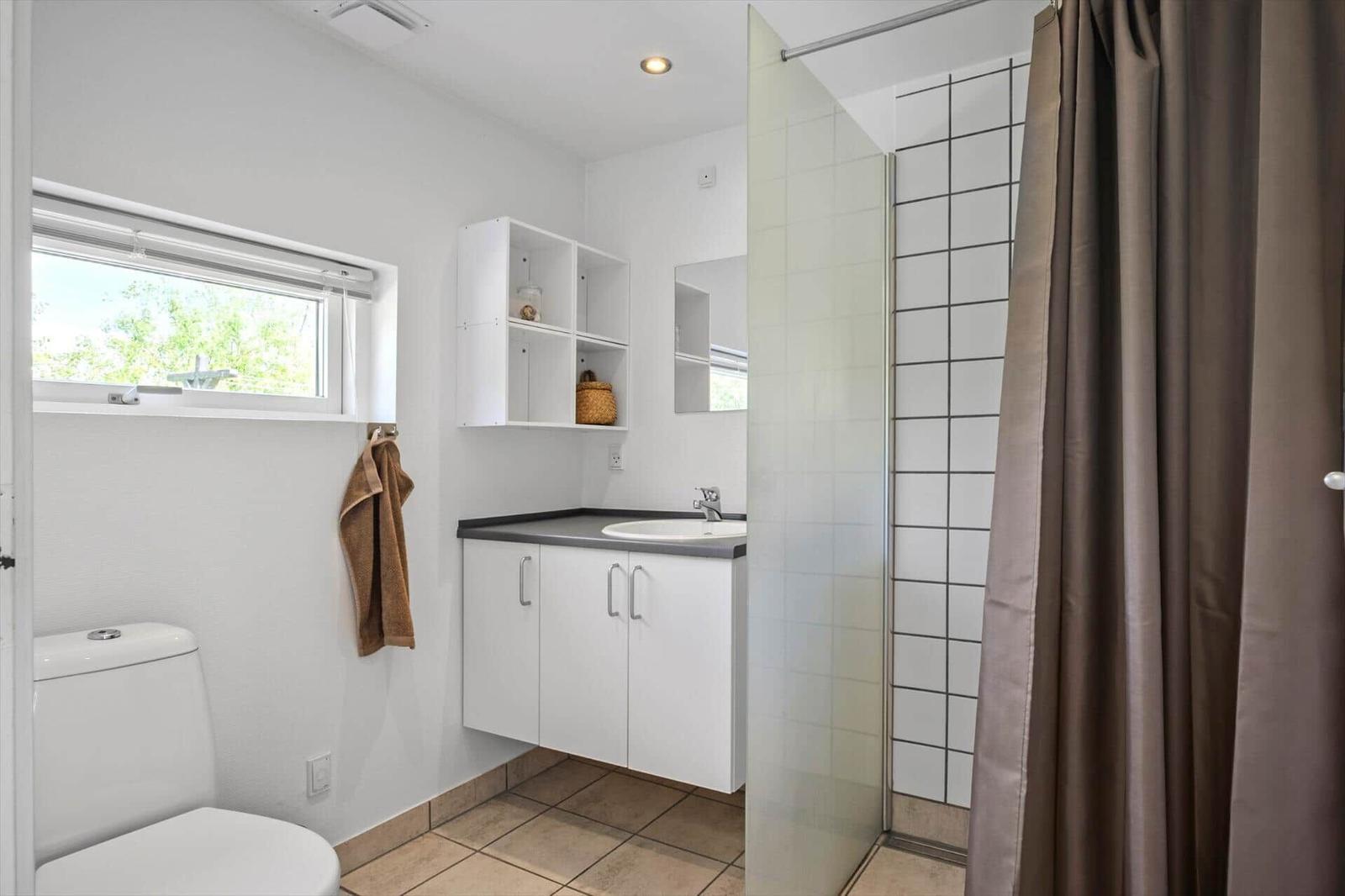 Bathroom with shower, sink, and toilet. Window, mirror, and storage.
