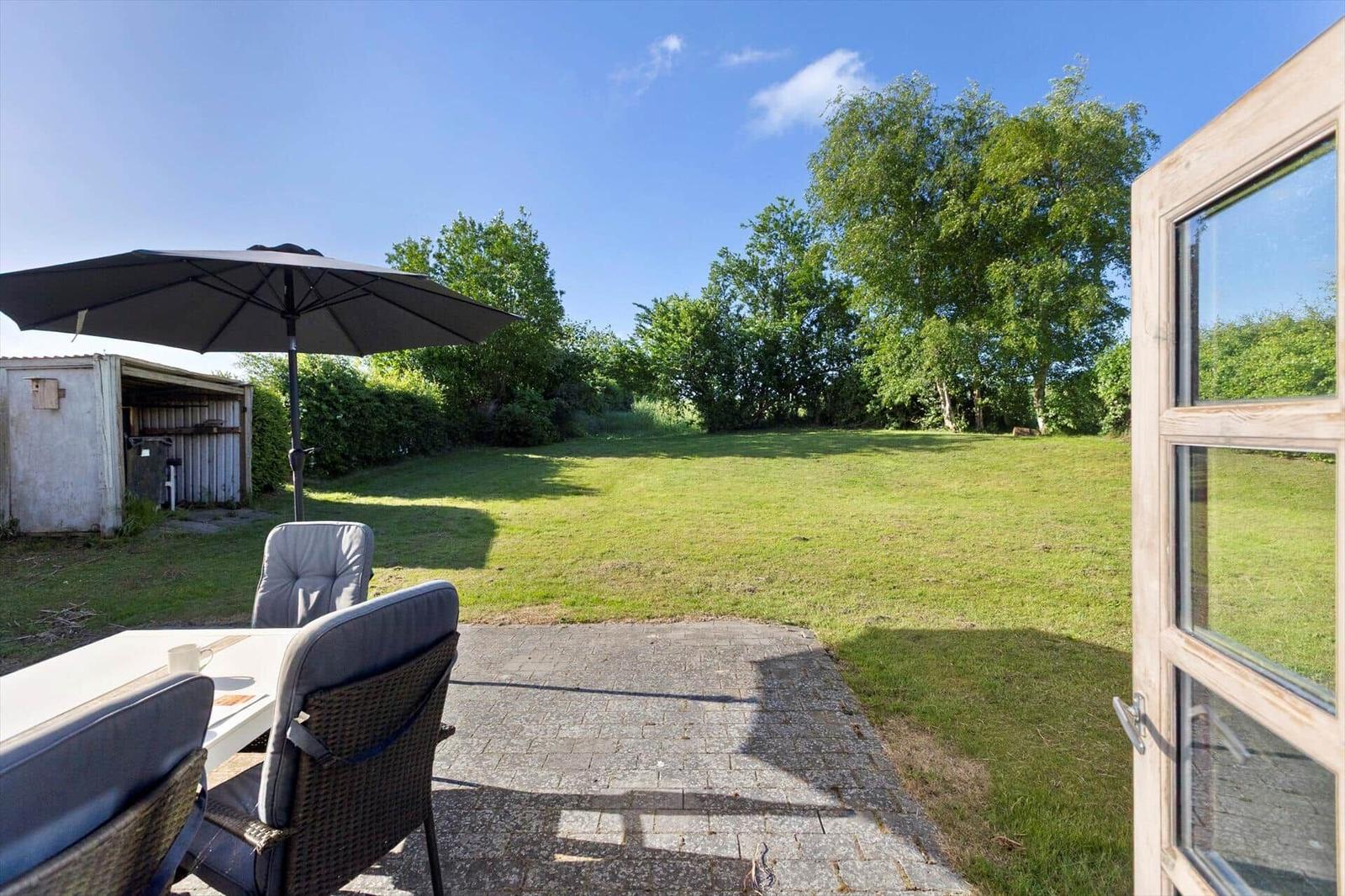Terrace with table, chairs, and umbrella. Lawn and trees in the background.