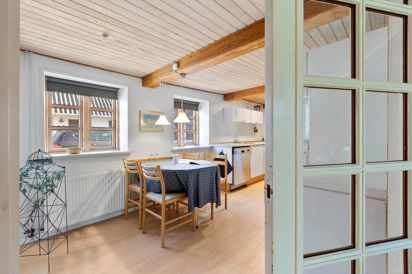 Dining room with table, chairs, and kitchen area. Wooden ceiling with beams and windows.