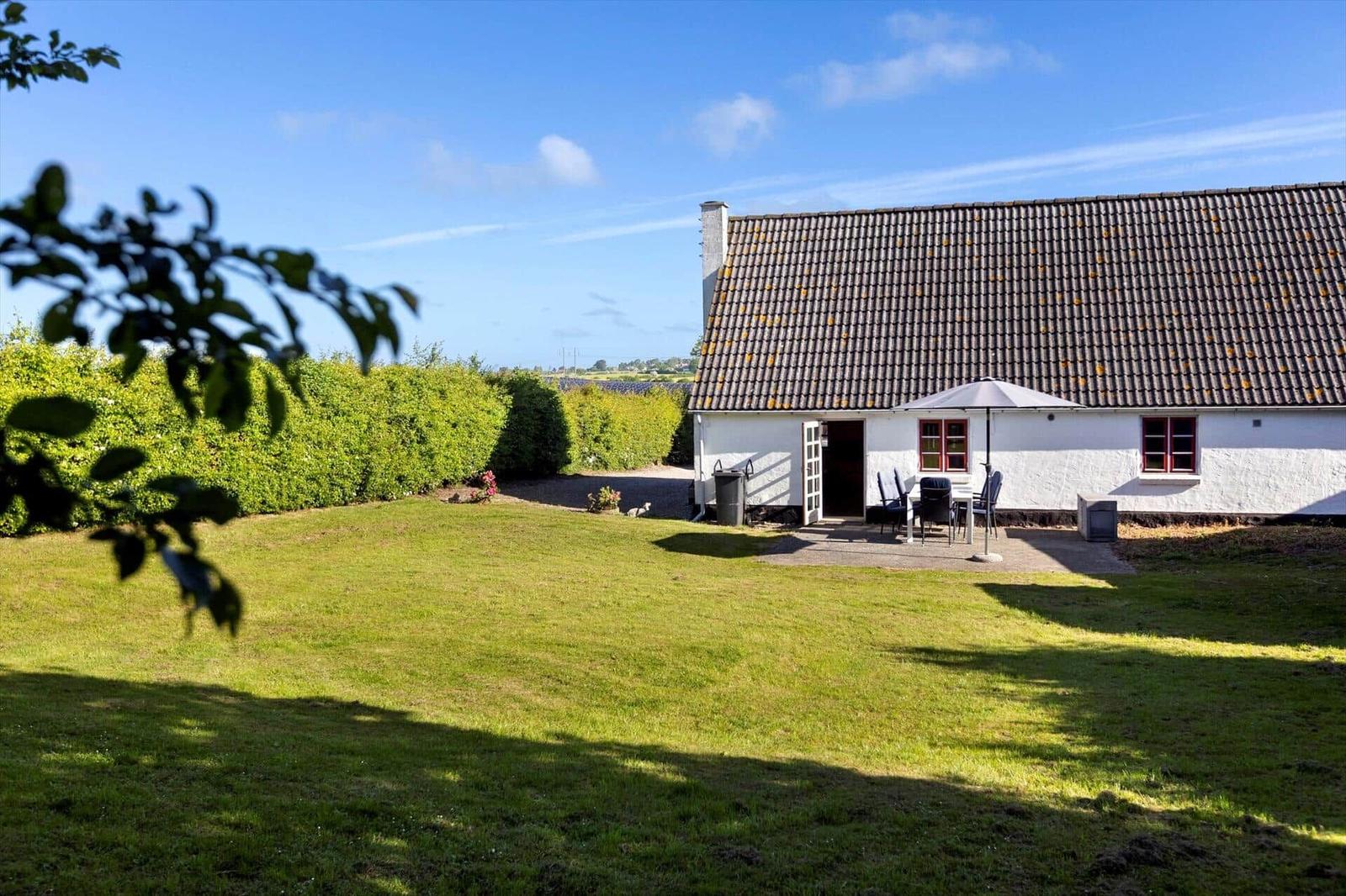 A white house with a brown roof, garden, and terrace with a table and chairs.