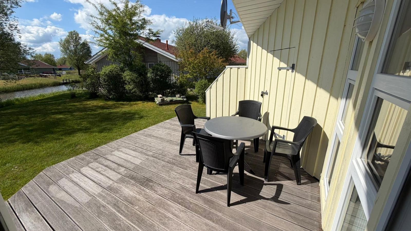 Terrasse mit Tisch und Stühlen neben gelbem Haus. Grünflächen und Wasser im Hintergrund.