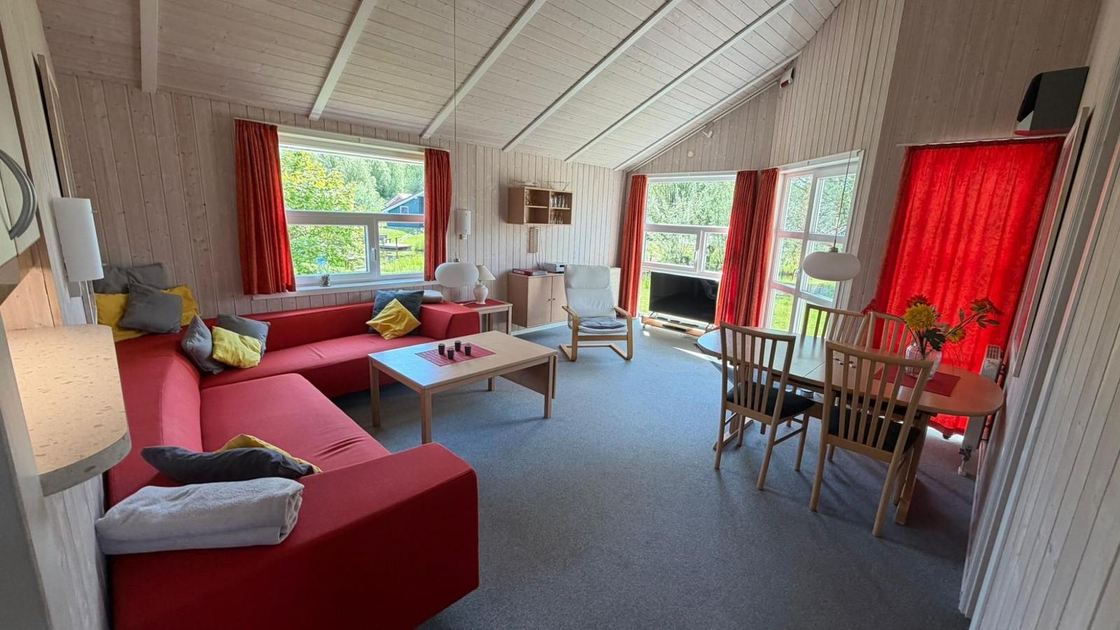 Wohn- und Essbereich mit rotem Sofa, Holztisch und TV. Rote Vorhänge und Holzwände.