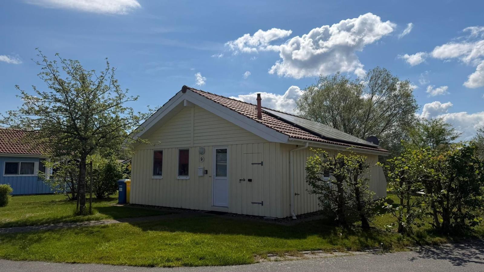 Ein gelbes Haus mit rotem Dach, umgeben von Grün und Bäumen unter blauem Himmel.