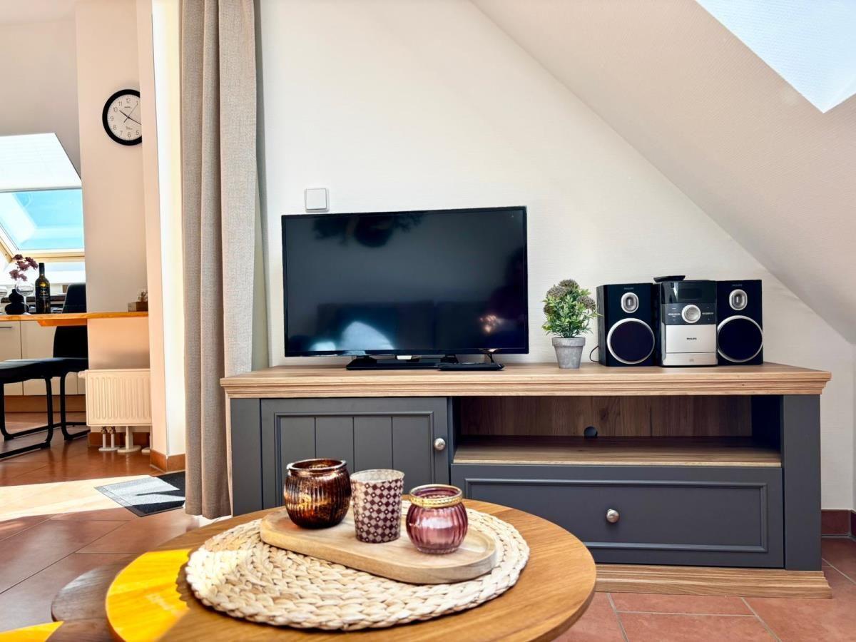 TV auf Holzboard mit Lautsprechern und Pflanze. Runder Tisch mit Kerzen und Korb. Sicht auf Küche.