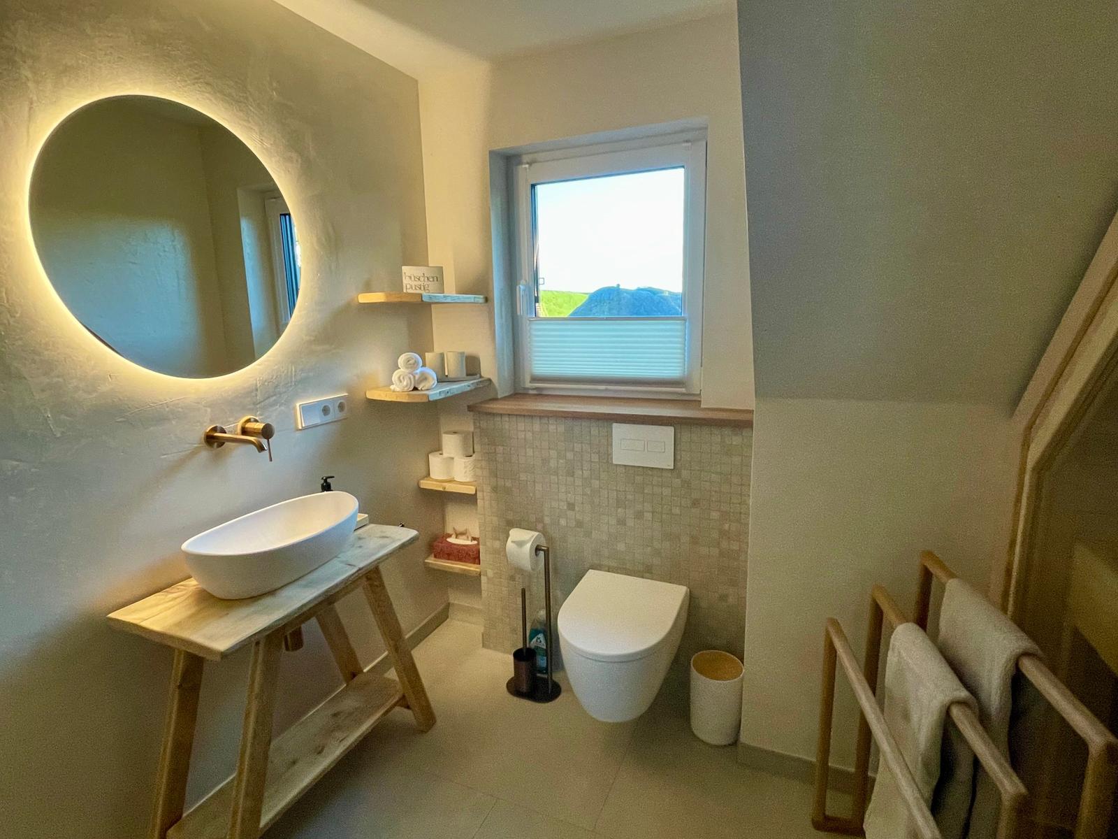 Badezimmer mit runden Spiegel, Waschtisch und Toilette. Fenster mit Blick auf Landschaft.