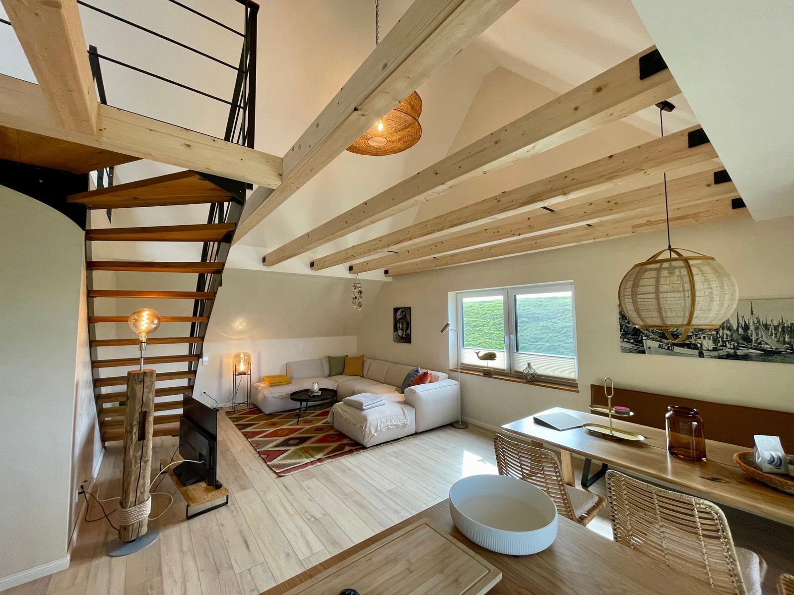 Wohnbereich mit Sofa, Tisch und Fenster. Holzdecken und Treppe sichtbar.