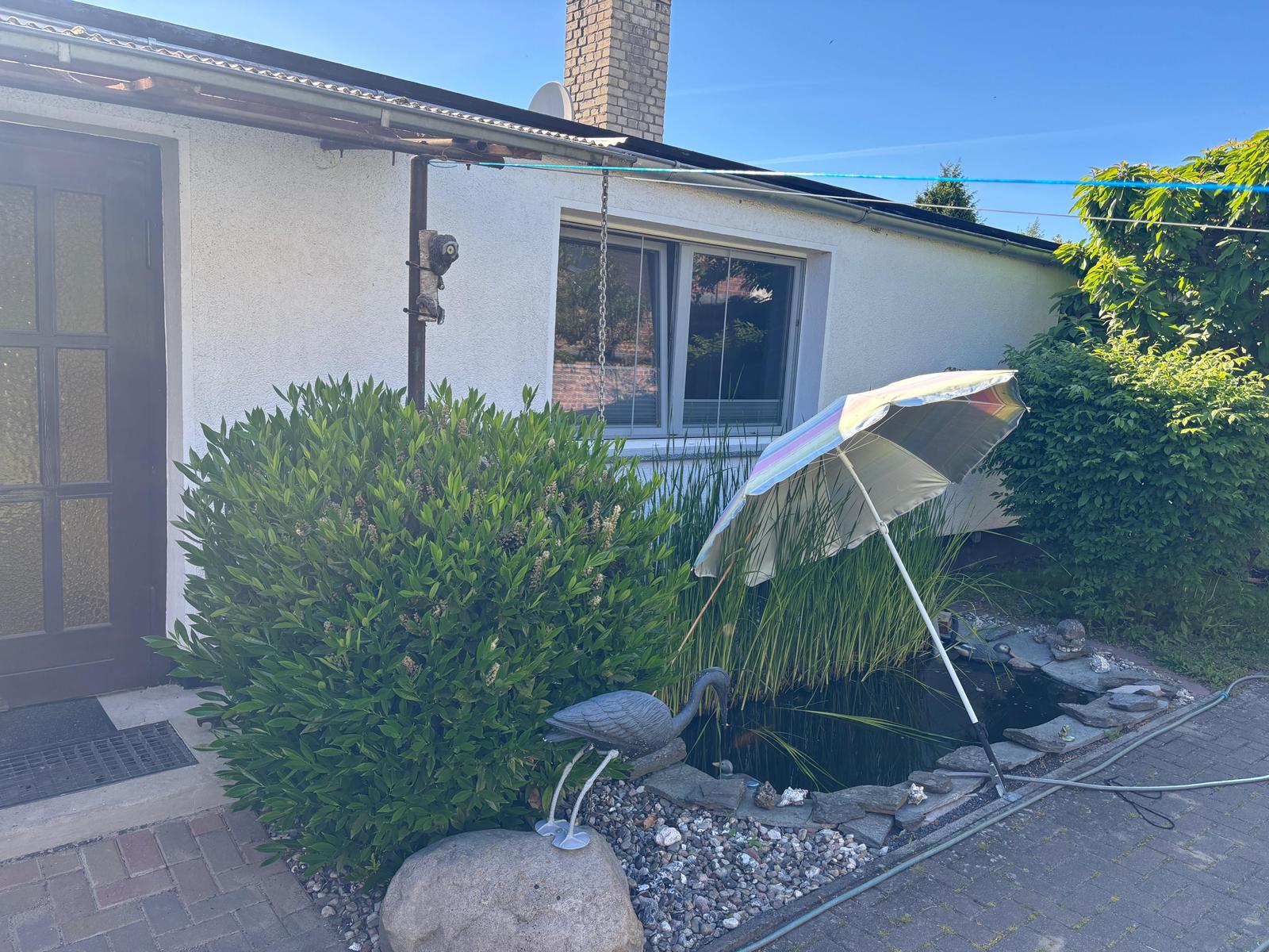 Haus mit Garten, Teich und Sonnenschirm. Pflanzen und Steinmauern umgeben den Außenbereich.