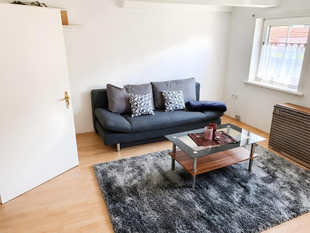 Ein Wohnzimmer mit grauem Sofa, Glas-Tisch und Holzboden. Fenster mit weißen Vorhängen.