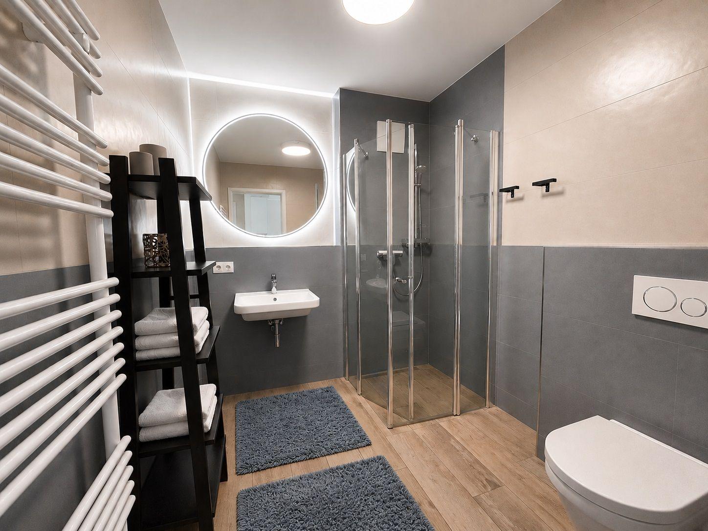 Badezimmer mit Dusche, Waschbecken und Toilette. Holzboden und graue Wandfliesen.
