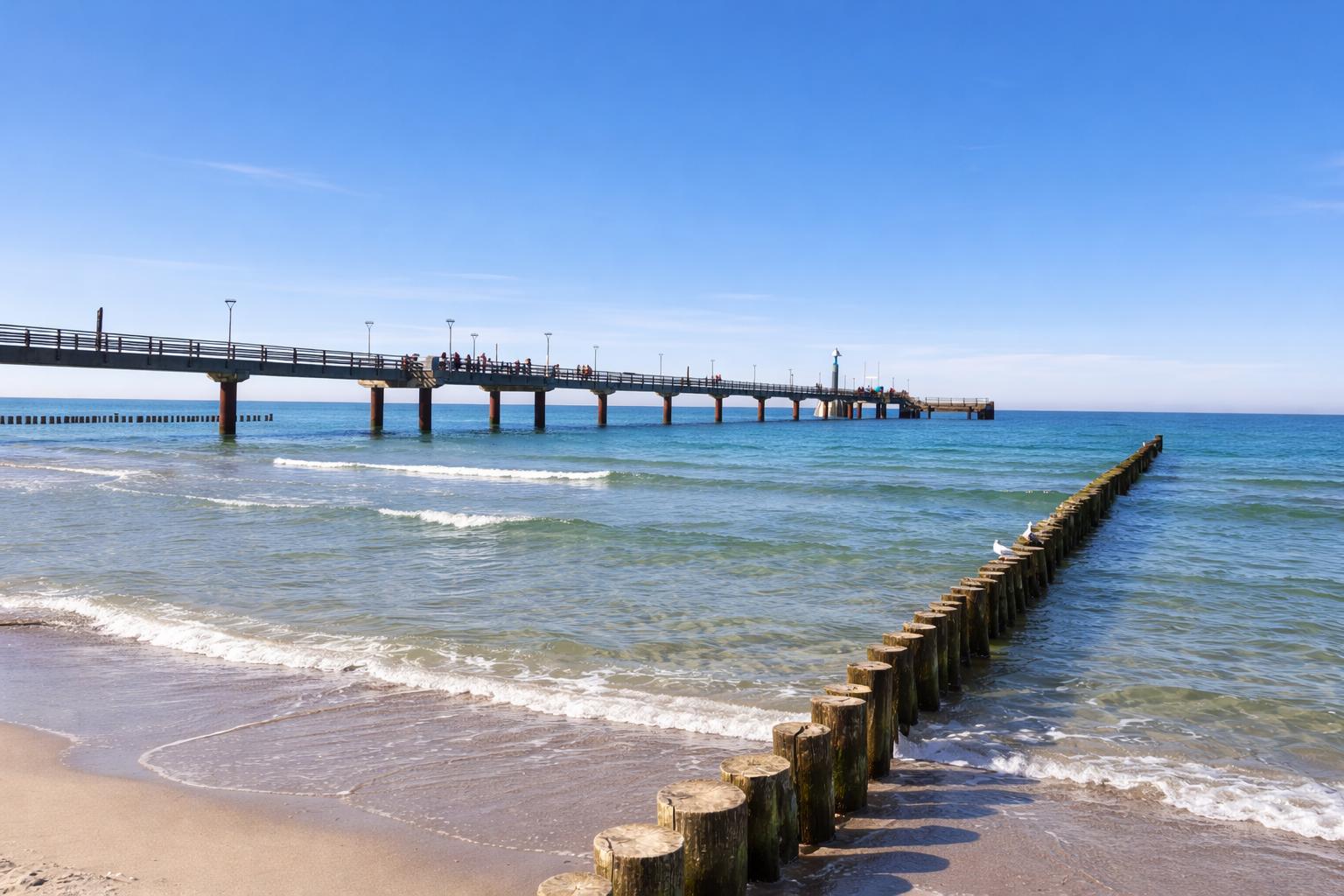 Langgezogener Steg führt über das Meer. Wellen schlagen an den Strand.