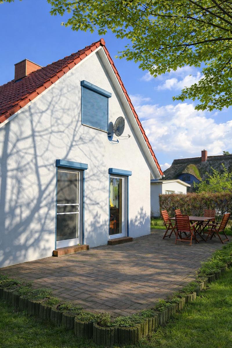 Wohnhaus mit Terrasse, Garten und Tisch mit Stühlen. Dach mit roten Ziegeln und blauen Fensterrahmen.
