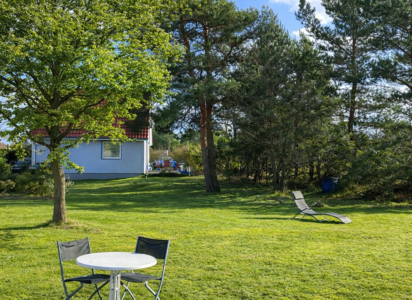 Grüner Garten mit Tisch, Stühlen und Liegestuhl. Im Hintergrund ein Haus mit rotem Dach.
