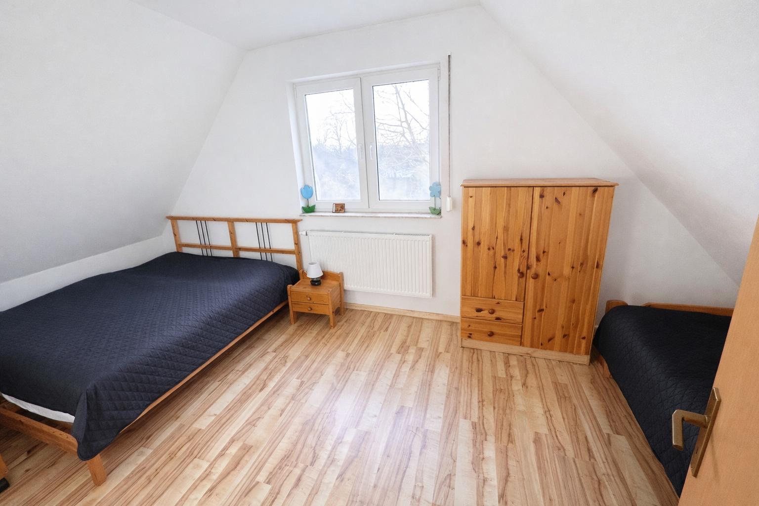 Schlafzimmer mit zwei Betten, Holzschrank und Fenster. Holzfußboden und weiße Wände.