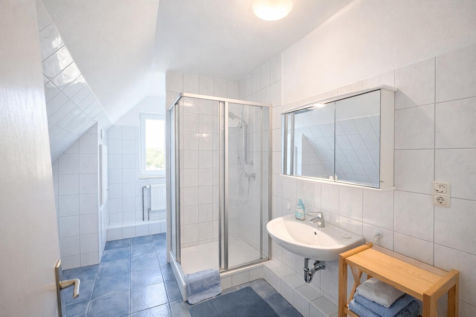 Badezimmer mit Dusche, Waschbecken und Spiegelschrank. Fliesenboden und Dachschrägen.