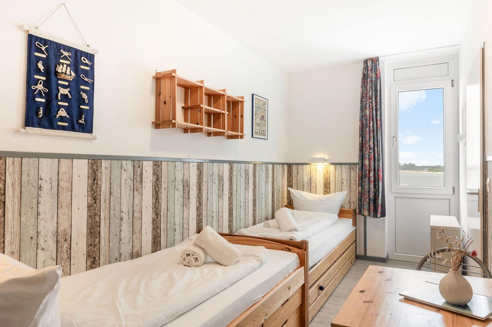Zwei Einzelbetten mit Holzgestell, Fenster mit Blick auf Wasser, Wanddekoration und Holzregal.