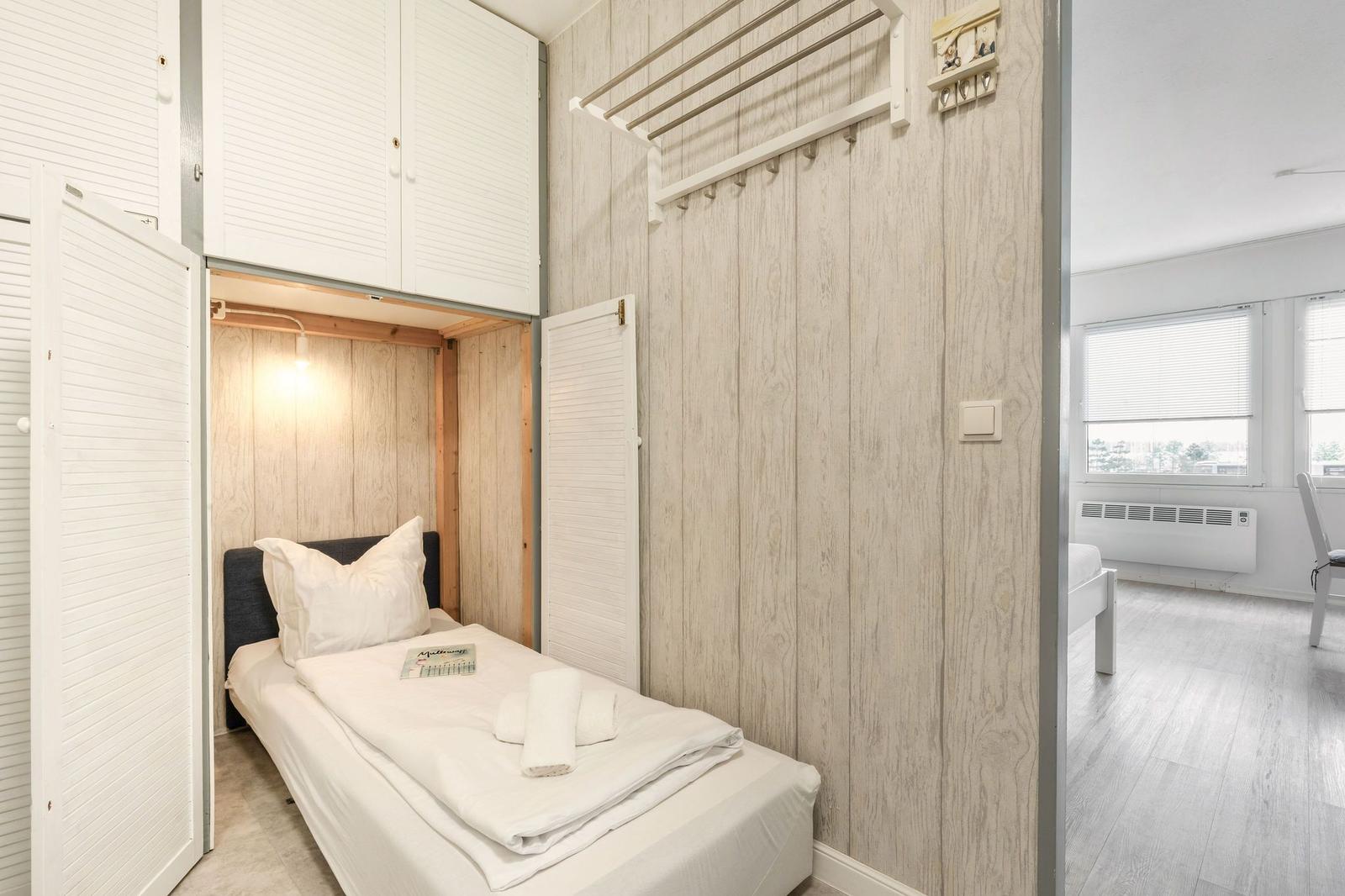 Schlafzimmer mit Bett, Kleiderhaken und Schrank. Durchgang zu Wohnbereich mit Fenster.
