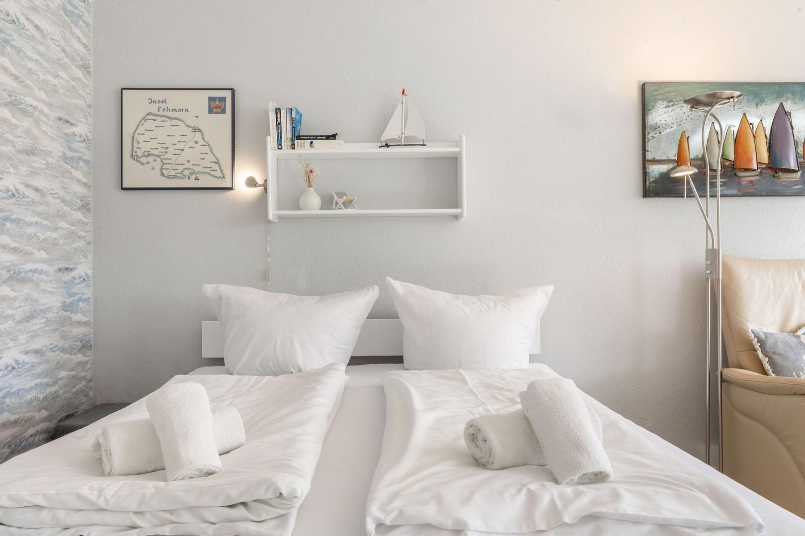 Schlafzimmer mit Doppelbett, weißen Kissen und Handtüchern. Wandbild und Regal mit Buchern.