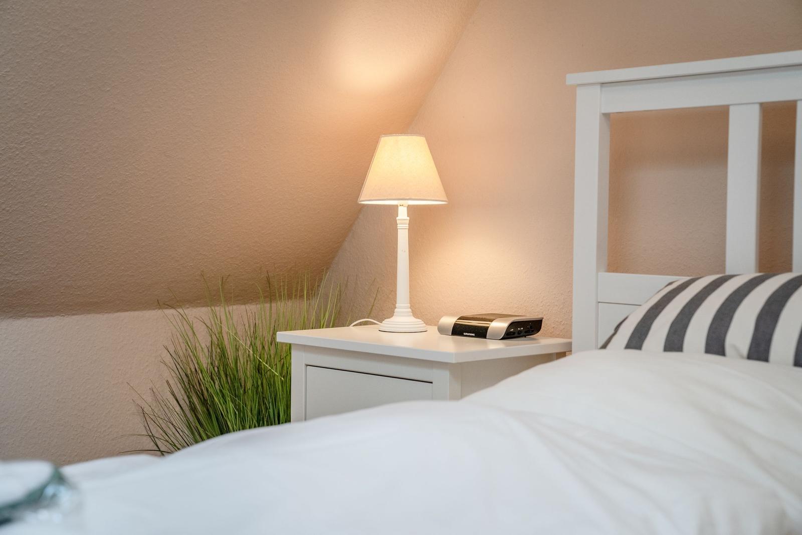 Bedroom with white bed, nightstand, and lamp. A plant hangs on the wall.