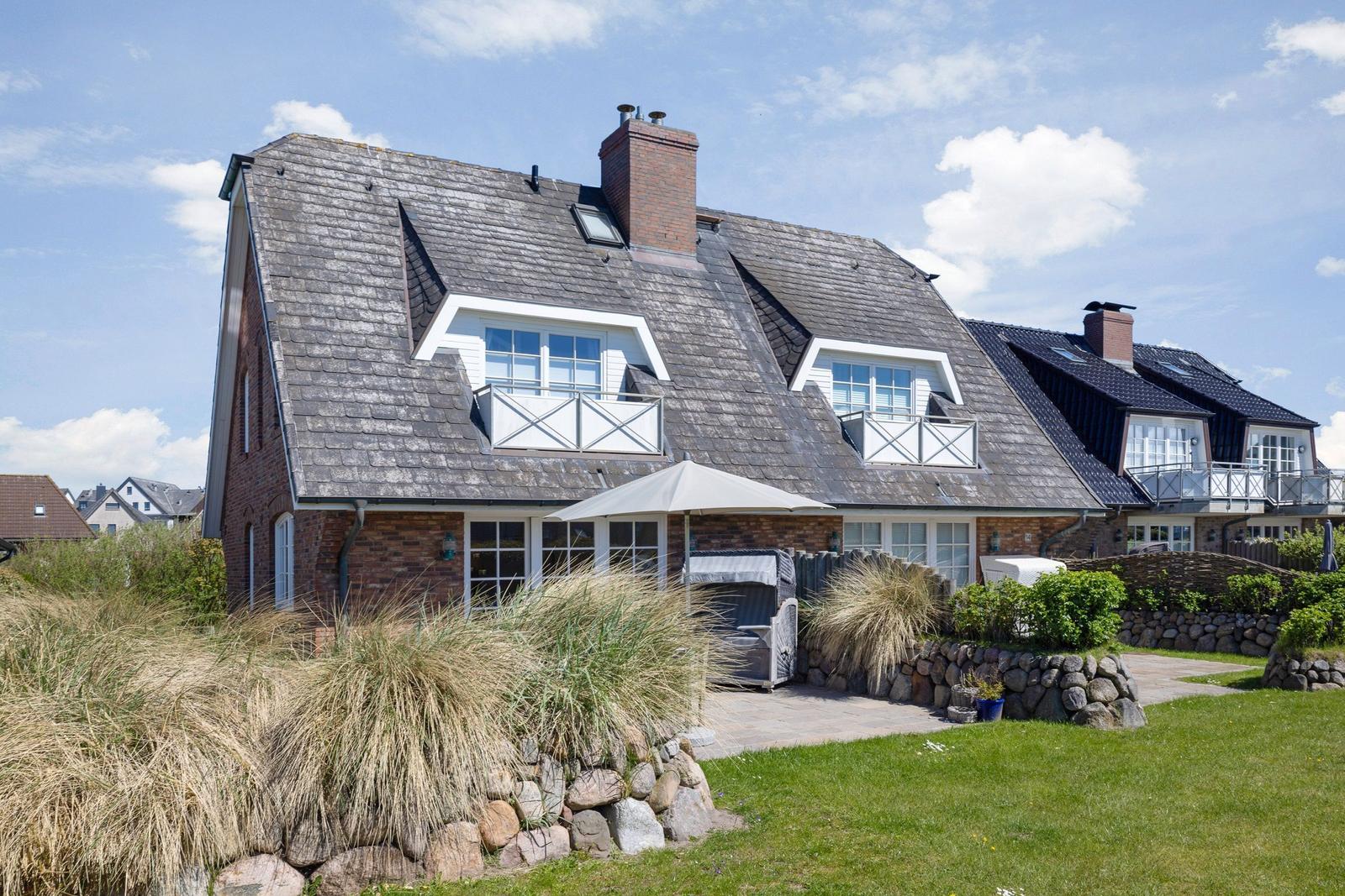 Haus mit Steinmauer, Gras und Terrasse unter blauem Himmel.