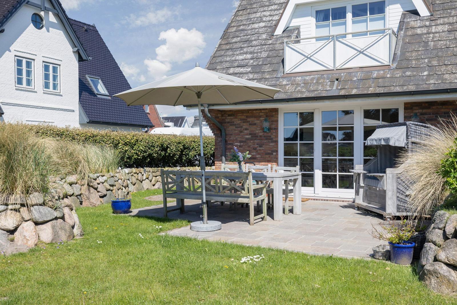 Terrasse mit Tisch, Bänken und Sonnenschirm. Hinterhof mit Gras, Steinmauer und Pflanzen.
